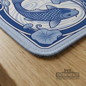 May include: A rectangular desk mat featuring a blue and white illustration of a koi fish swimming among stylized water ripples and lotus leaves. The mat has stitched edges and includes the text DESKNEKO and WWW.DESKNEKO.COM in the corner.