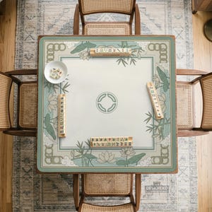 May include: Overhead view of a square mahjong table with a floral and bamboo design in shades of green and white. The table is surrounded by four wooden chairs with woven backs. Mahjong tiles are placed on the table.