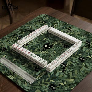 May include: A Mahjong set arranged on a green mat with a leaf and black cat pattern. The white tiles are arranged in a square, with Chinese characters and symbols visible. A clear plastic tile holder is in the foreground.