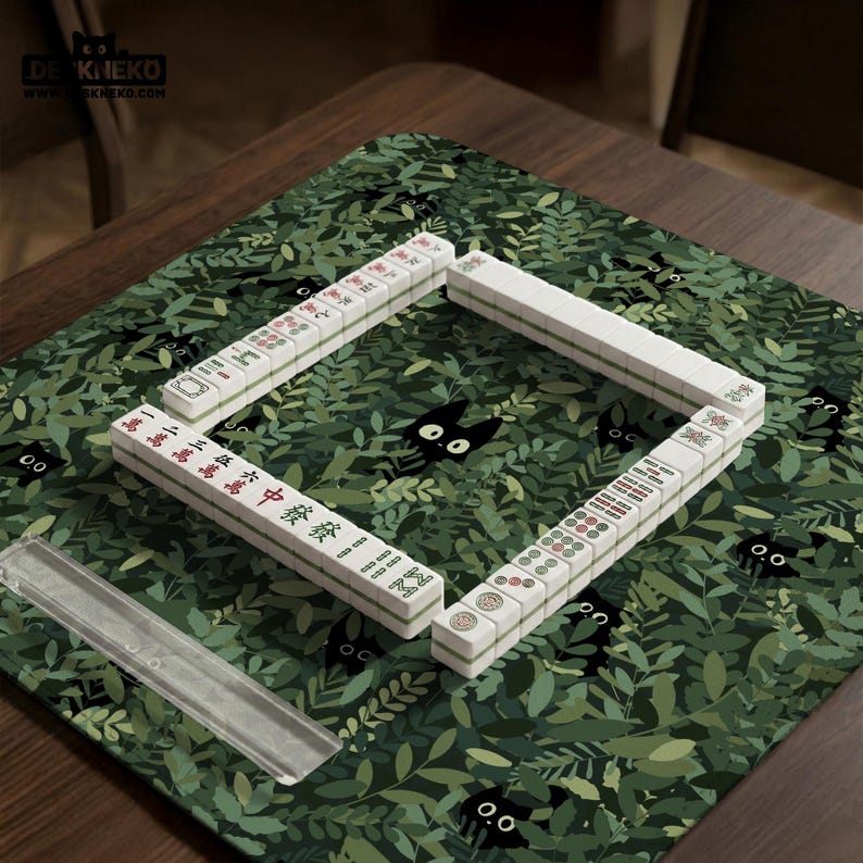 May include: A Mahjong set arranged on a green mat with a leaf and black cat pattern. The white tiles are arranged in a square, with Chinese characters and symbols visible. A clear plastic tile holder is in the foreground.