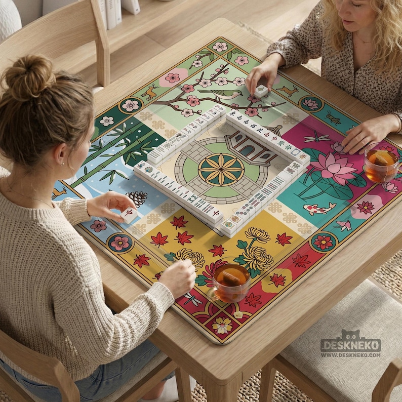 May include: A square wooden table with a colorful Mahjong game mat featuring floral and nature-themed panels. Two people are playing Mahjong, with tiles and tea cups visible. The mat has a variety of colors and patterns.