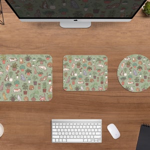 Puede incluir: Una configuraci&oacute;n de escritorio de computadora con un monitor, teclado, rat&oacute;n y accesorios de escritorio a juego. Los accesorios incluyen una alfombrilla de rat&oacute;n cuadrada, un tapete de escritorio rectangular y un posavasos redondo, todos con un patr&oacute;n repetido de gatos y plantas en macetas sobre un fondo verde.