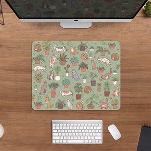 Puede incluir: Alfombrilla de rat&oacute;n rectangular con fondo verde claro. Presenta un patr&oacute;n repetido de plantas en macetas e ilustraciones de gatos en varias poses. Los gatos son de color marr&oacute;n, gris y naranja.