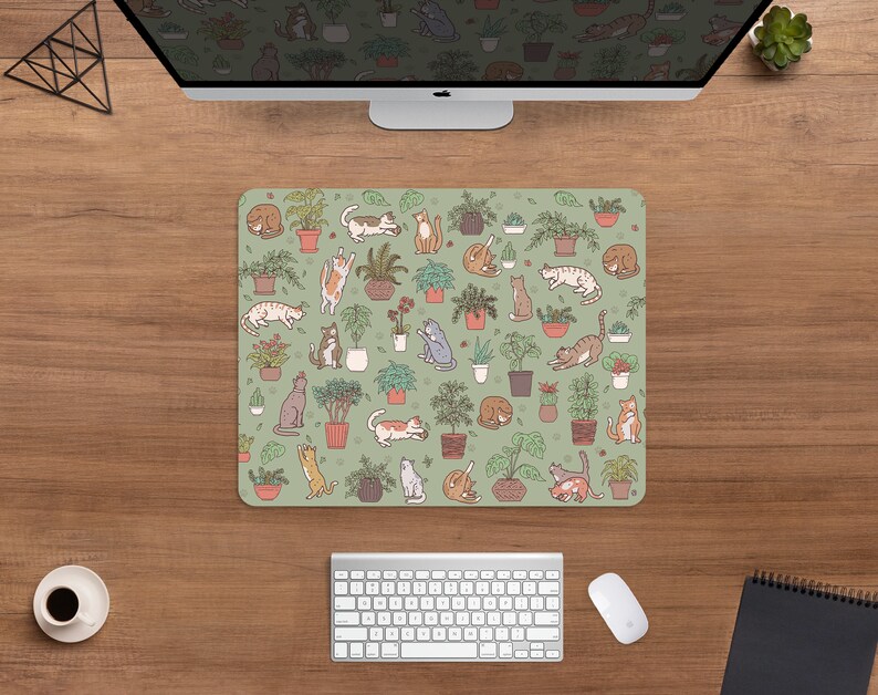 Puede incluir: Alfombrilla de rat&oacute;n rectangular con fondo verde claro. Presenta un patr&oacute;n repetido de plantas en macetas e ilustraciones de gatos en varias poses. Los gatos son de color marr&oacute;n, gris y naranja.