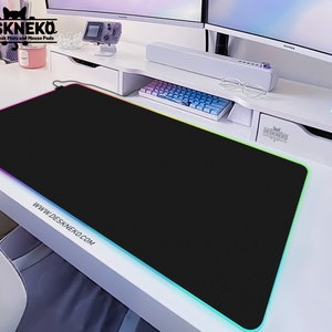 Op de afbeelding: Een zwarte rechthoekige bureaumat met een kleurrijke LED-rand. De mat ligt op een witte bureau met een toetsenbord en monitor op de achtergrond. De tekst "DESKNEKO Aesthetic Desk Mats and Mouse Pads" is zichtbaar.