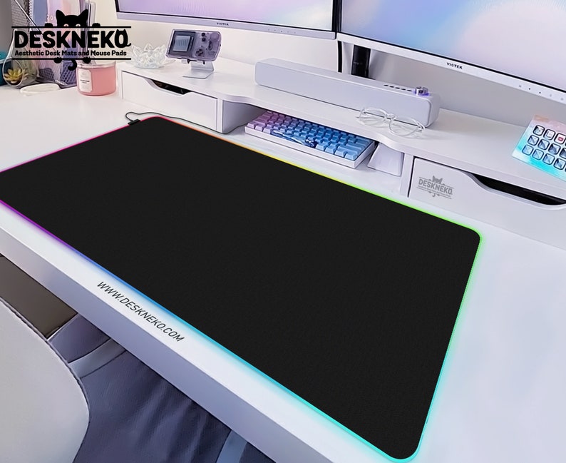 Op de afbeelding: Een zwarte rechthoekige bureaumat met een kleurrijke LED-rand. De mat ligt op een witte bureau met een toetsenbord en monitor op de achtergrond. De tekst "DESKNEKO Aesthetic Desk Mats and Mouse Pads" is zichtbaar.