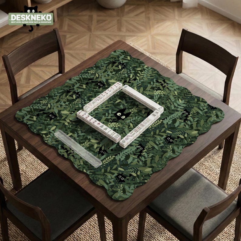 May include: A square, dark green placemat with a leaf pattern and black cat silhouettes. The placemat is on a dark wood table with a white and black game board. The text "DESKNEKO" is in the upper left corner.