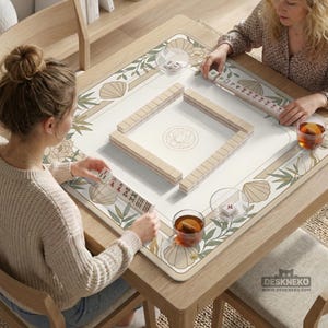 May include: A wooden mahjong table with a cream-colored playing surface featuring a floral and seashell design. The table is set for a game, with tiles arranged and players holding tiles. The table has a decorative border and is surrounded by chairs.