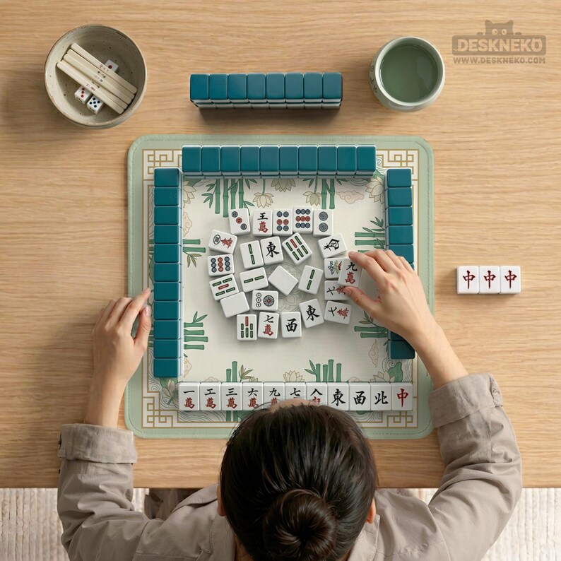 Puede incluir: Una persona jugando al Mahjong. El juego incluye fichas turquesas, dados y un tapete con tem&aacute;tica de bamb&uacute;. Las fichas son blancas con s&iacute;mbolos rojos y negros. Un cuenco peque&ntilde;o y una taza de t&eacute; tambi&eacute;n est&aacute;n sobre la mesa de madera.