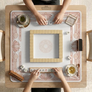 May include: Overhead shot of a mahjong game in progress. The wooden table features a floral design in pink and white. Beige mahjong tiles are arranged in a square, with players holding tiles. Teacups and accessories are also on the table.