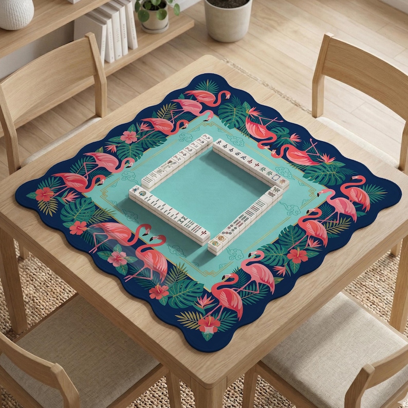 May include: A square Mahjong game mat with a teal center and a navy blue border. The border features pink flamingos, tropical flowers, and green leaves. The mat is on a wooden table with a set of Mahjong tiles.