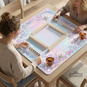 May include: A wooden mahjong table with a pastel-colored mat featuring bows, seahorses, and shells. Two people are playing mahjong, with tiles arranged in a square. Tea cups and tile racks are also visible.