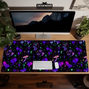 May include: A black desk mat featuring a vibrant pattern of purple and pink mushrooms, moons, stars, and other mystical symbols. The mat has the text "DESKNEKO" and "WWW.DESKNEKO.COM" printed on it. A white keyboard and mouse are on the mat.