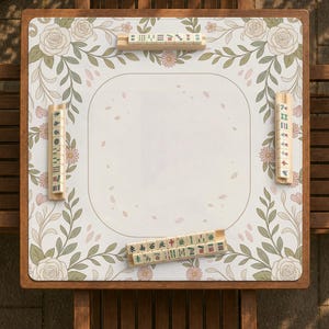 May include: A square mahjong mat featuring a white center with scattered pink petal illustrations, bordered by a floral pattern of roses, leaves, and small daisies. Four wooden racks hold mahjong tiles with various characters and symbols.