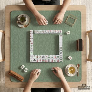 以下が含まれることがあります： 木製のテーブルで進行中の麻雀ゲームの俯瞰図。緑色のマットは波模様です。白いタイルは正方形に配置され、プレイヤーの手が見えます。ティーカップと木製のタイルホルダーも存在します。