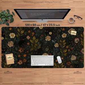 Op de afbeelding: Een zwarte bureaumat met een bloemen- en kattenontwerp. De mat meet 120 x 60 cm en heeft een patroon van bloemen, bladeren en verborgen zwarte kattengezichten. Een toetsenbord, muis en monitor liggen op de mat.
