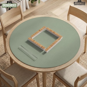 May include: A round sage green desk mat featuring a textured surface, placed on a wooden table. The mat displays a square arrangement of light brown wooden tiles and a clear rectangular acrylic tile holder. The logo DESKNEKO is visible.