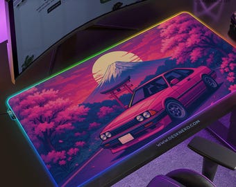 May include: Gaming mouse pad with a vibrant Japanese-inspired design. Features a pink car, Mount Fuji, and cherry blossoms against a sunset backdrop. The pad has a colorful LED edge and the text "DESKNEKO".