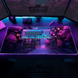 May include: A rectangular DESKNEKO mouse mat featuring a Japanese street scene with pink cherry blossom trees and buildings. A backlit keyboard sits on the mat. The mat has a glowing purple edge. The website address www.deskneko.com is visible.