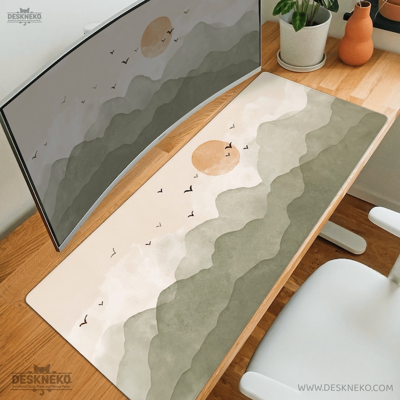 May include: A rectangular desk mat with a landscape design featuring a sun, mountains, and birds in shades of beige, green, and brown. The mat is placed on a wooden desk, with a computer monitor and a white chair visible.