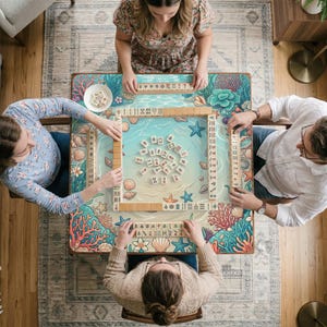 May include: A group of four people playing Mahjong on a table with a beach-themed design. The table features a light blue ocean scene with seashells, starfish, and coral. The game pieces are wooden, and the table is surrounded by players.