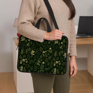 May include: A black laptop bag with a floral pattern in shades of green and cream is carried over the shoulder. The bag has a black handle and a black shoulder strap. The floral design features various leaves and flowers.