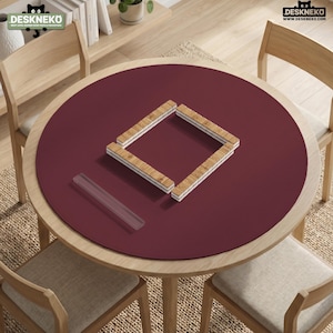 May include: A circular, deep burgundy desk mat centered on a round wooden table. The mat features a square arrangement of light-colored, rectangular wooden tiles and a single, clear, rectangular acrylic block resting on its smooth surface.