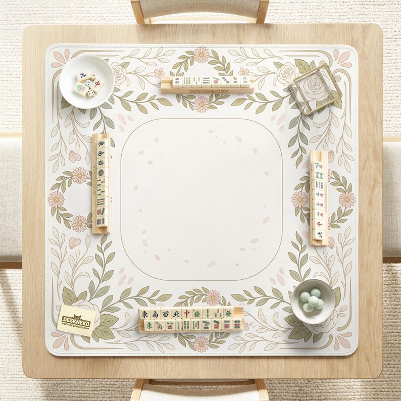 May include: A square mahjong mat featuring a cream-colored center with a floral border of green leaves and pink flowers. The mat includes four wooden tile racks, a white bowl with mahjong tiles, a small bowl with green glass beads, and a logo card.