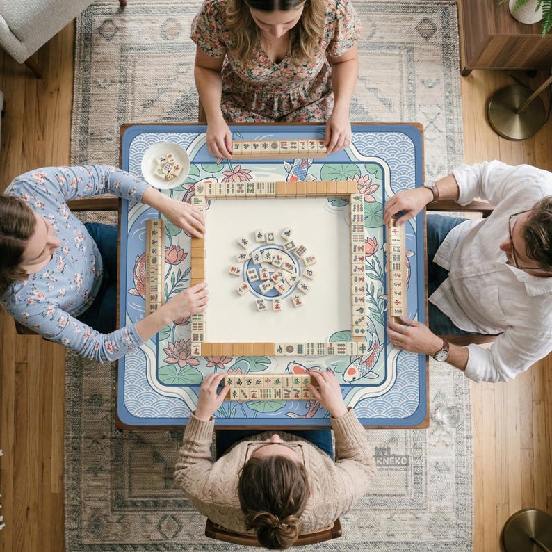 May include: An overhead shot of a Mahjong game in progress. The blue table has a floral and koi carp design. The beige game tiles have black symbols. Four people are seated around the table, each holding tiles.