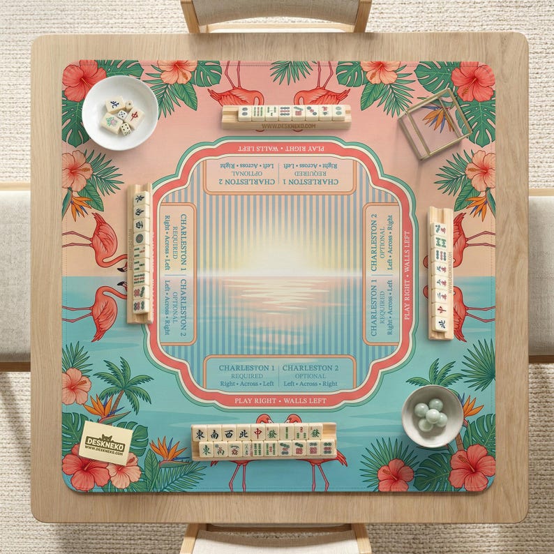 May include: A square mahjong mat featuring a tropical flamingo and hibiscus flower pattern in pink, green, and blue. The center includes game instructions and Charleston rules. Four wooden tile racks, a white bowl with tiles, and a bowl with dice are present.