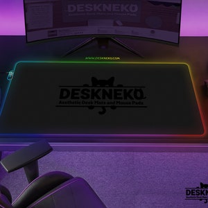 Op de afbeelding: Een zwart rechthoekig bureau-onderlegger met een oplichtende regenboogrand. De mat toont de tekst "DESKNEKO Aesthetic Desk Mats and Mouse Pads" en het websiteadres "WWW.DESKNEKO.COM". Een computermonitor, muis en gamecontroller zijn ook zichtbaar.