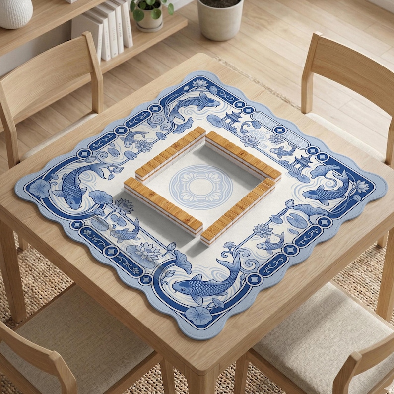 May include: A square mahjong mat with a scalloped edge featuring a blue and white pattern of koi fish, lotus flowers, and pagodas. In the center, four wooden mahjong racks are arranged in a square formation on a white surface with a circular design.