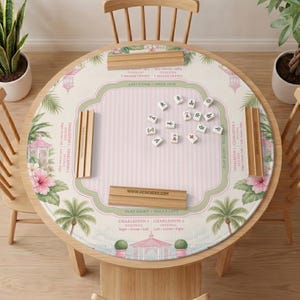 May include: A round Mahjong table mat featuring a light pink striped center, green borders, and tropical illustrations of pink hibiscus flowers, palm trees, and gazebos. Four wooden tile racks are placed on the mat with Mahjong tiles in the center.