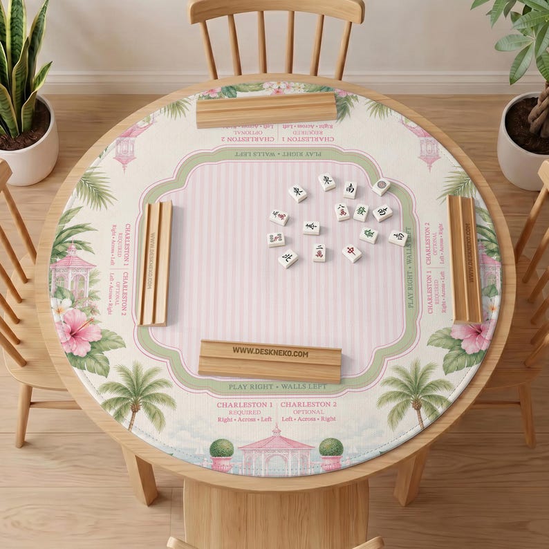 May include: A round Mahjong table mat featuring a light pink striped center, green borders, and tropical illustrations of pink hibiscus flowers, palm trees, and gazebos. Four wooden tile racks are placed on the mat with Mahjong tiles in the center.