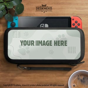 Può includere: Una custodia nera con la scritta "YOUR IMAGE HERE" e impronte di zampe, che contiene una console di gioco verde acqua e rossa. La custodia è su una superficie di legno, con una piccola pianta e una tazza di caffè sullo sfondo. La custodia ha un cinturino da polso.