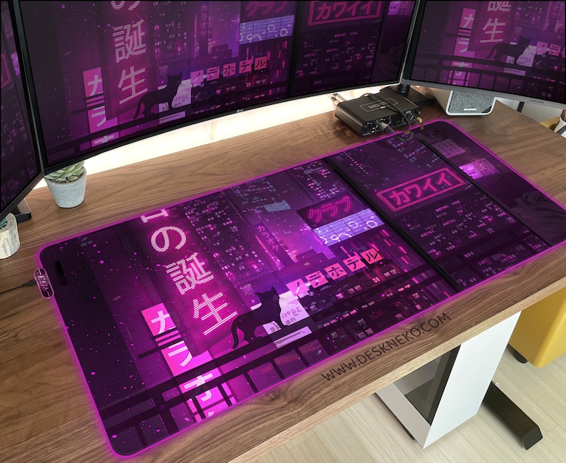 Op de afbeelding: Een rechthoekige muismat met een neonpaars stadsgezichtontwerp. De mat heeft Japanse tekst en een silhouet van een kat. De woorden "KAWAII" en "DESKNEKO.COM" zijn ook zichtbaar. De mat heeft een gloeiende paarse rand.