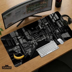 May include: Black and white desk mat featuring a nighttime cityscape of Tokyo with neon signs and a crescent moon. The mat has the text "TOKYO" and "DESKNEKO" printed on it. A banana, keyboard, mouse, and headphones are on the desk.