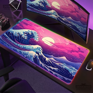 Puede incluir: Alfombrilla de ratón rectangular para juegos con un diseño de ola japonesa estilizada en tonos azules, rosas y blancos. La alfombrilla tiene un borde LED colorido y el texto "DESKNEKO" está impreso en el borde inferior.
