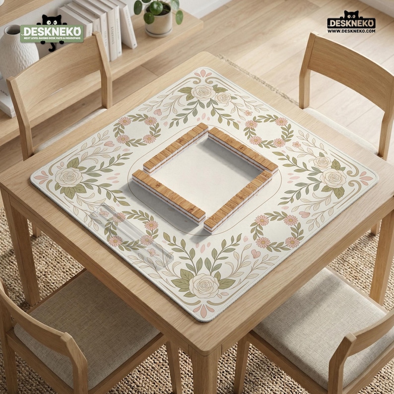 May include: A square mahjong mat featuring a cream-colored surface with a floral border of green leaves, pink roses, and small orange flowers. In the center, a wooden frame creates a square boundary. The mat displays the DESKNEKO logo.
