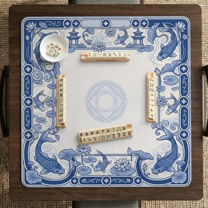 May include: A square mahjong mat featuring a blue and white design with koi fish, lotus flowers, and pagodas. The mat includes four wooden tile racks holding mahjong tiles and a small white bowl containing several dice on the surface.