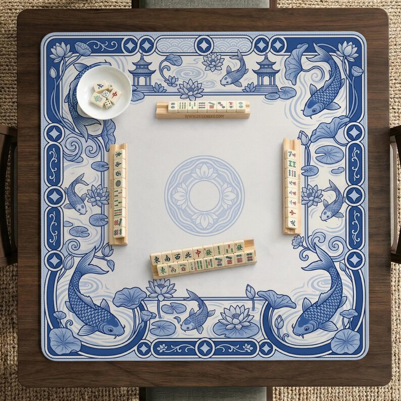 May include: A square mahjong mat featuring a blue and white design with koi fish, lotus flowers, and pagodas. The mat includes four wooden tile racks holding mahjong tiles and a small white bowl containing several dice on the surface.