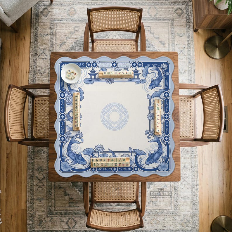May include: A square mahjong table mat featuring a blue and white pattern with koi fish, lotus flowers, and pagoda motifs. The mat has a scalloped edge and is displayed on a wooden table with four cane-back chairs, with mahjong tiles placed on top.