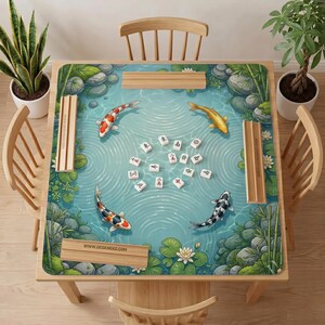 May include: A square mahjong mat featuring a pond scene with four koi fish, lily pads, and rocks. The center displays scattered white mahjong tiles with black and red characters. Four wooden tile racks are positioned along the edges of the mat.