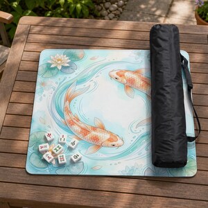 May include: A square mahjong mat featuring a light blue watercolor background with two orange koi fish swimming in a circle, surrounded by white lotus flowers and green lily pads, accompanied by a black carrying case and several mahjong tiles.