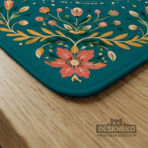 May include: A rectangular desk mat with a teal background featuring a symmetrical floral pattern in shades of coral, peach, and yellow. The design includes stylized flowers, leaves, and small berries. The mat has stitched edges and a logo.