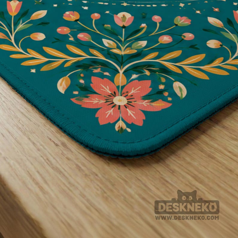 May include: A rectangular desk mat with a teal background featuring a symmetrical floral pattern in shades of coral, peach, and yellow. The design includes stylized flowers, leaves, and small berries. The mat has stitched edges and a logo.