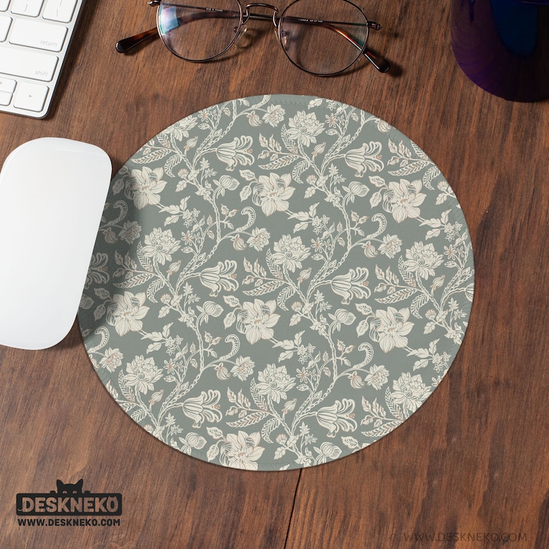 Puede incluir: Alfombrilla de rat&oacute;n redonda con un estampado floral en crema y marr&oacute;n claro sobre un fondo verde apagado. La alfombrilla est&aacute; sobre un escritorio de madera junto a un rat&oacute;n y un teclado blancos. La marca "DESKNEKO" est&aacute; impresa en la parte inferior izquierda.