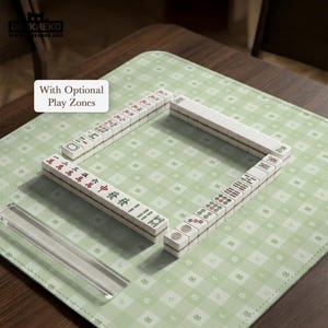 May include: A Mahjong set arranged on a light green play mat with a frog pattern. The white tiles have red, green, and black symbols. The text "With Optional Play Zones" is in a white box. A clear acrylic piece is in the foreground.