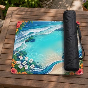May include: A square mahjong mat featuring a vibrant tropical beach scene with turquoise water, white waves, and colorful hibiscus flowers. The mat includes a black carrying case with a shoulder strap and several white mahjong tiles.