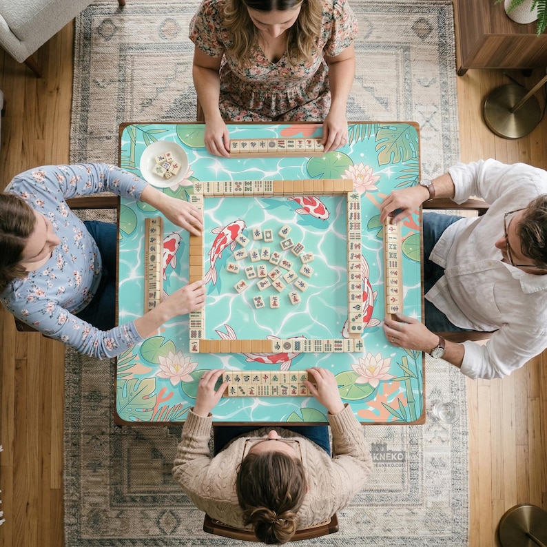 May include: A square mahjong table with a light blue surface featuring a koi fish, lotus flower, and tropical leaf pattern. The table is set with four wooden tile racks and a central collection of mahjong tiles arranged for a game.